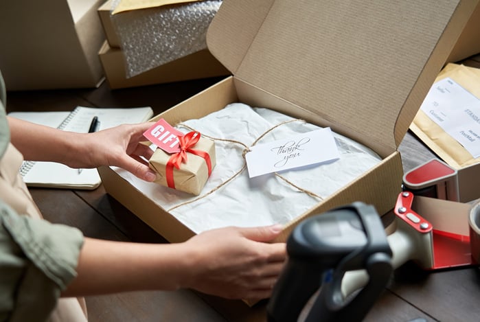 Mercado Pago: pessoa montando um pacote de itens da sua loja vendido para as festas de fim de ano em uma caixa de papelão, onde tem um tecido e um brinde. 