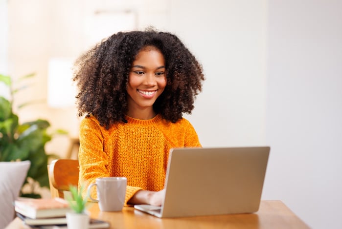 Mercado Pago: mulher sentada à mesa, mexendo no seu notebook para fazer a gestão do Pagamento Recorrente Mercado Pago do seu e-commerce. Ela está sorrindo e, na mesa, tem uma xícara e alguns cadernos.