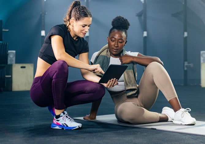 Mercado Pago: mulher em uma academia ao lado da sua personal trainer, que está lhe mostrando a ficha de exercícios em um tablet.