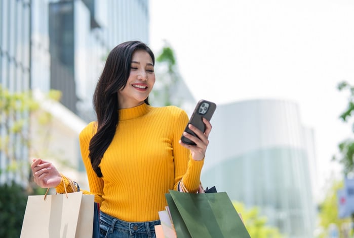Mercado Pago: mulher sorrindo, segurando sacolas nos braços e um celular em uma das mãos, enquanto finaliza mais compras usando vale-presente.