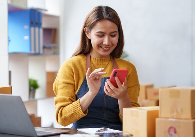 Mercado Pago: vendedora com o celular na mão, notebook na mesa e caixas espalhadas pelo espaço enquanto ela calcula a taxa de conversão de vendas com um sorriso no rosto.