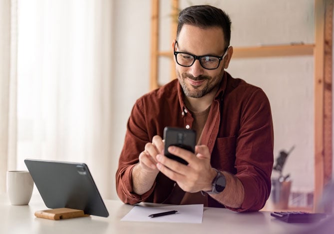 Mercado Pago: imagem de um empresário sentado à sua mesa de trabalho, enquanto pesquisa em seu celular quais recursos tecnológicos podem ajudar a vender mais em sua loja e a melhorar o relacionamento com o cliente. O empresário usa uma camisa de manga longa terracota e óculos e, sobre a mesa, há uma folha em branco com uma caneta, além de um tablet apoiado em um suporte e uma caneca branca.