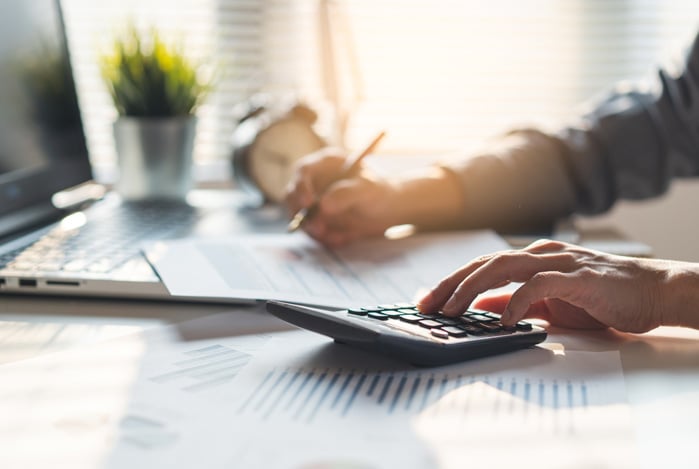 Mercado Pago: mãos de um homem apoiadas em uma mesa com computador, papéis e uma calculadora. Com a mão esquerda, ele faz cálculos e com a direita, anota as estratégias de redução de custos da sua empresa em um papel.