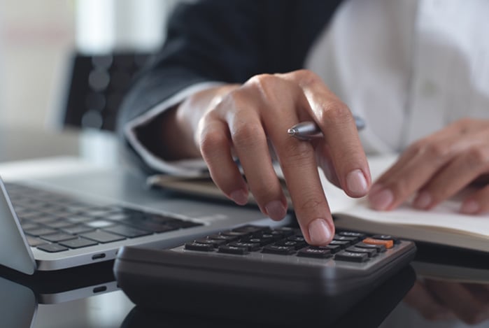 Mercado Pago: mãos digitando em uma calculadora apoiada em uma mesa, onde também há um notebook e um caderno, para a pessoa estudar sobre liquidez diária nos seus investimentos.