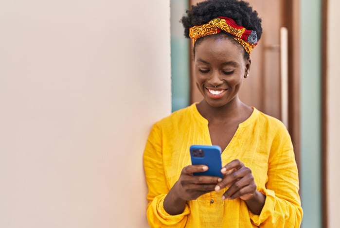 Mulher negra com cabelo crespo arrumado com um lenço estampado, vestindo blusa amarela e segurando um celular enquanto sorri devido ao sucesso em fazer o seu planejamento anual no app Mercado Pago. 